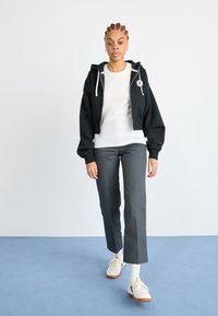 Black cropped hoodie with white drawstrings, worn over a white ribbed shirt, paired with grey straight-leg trousers and cream sneakers.