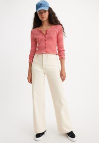 Cardigan rayé rouge et blanc, décolleté en V, coupe ajustée, associé à un pantalon large taille haute crème et des baskets basses noires. Casquette bleue en accent.