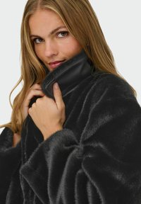 Donna con lunghi capelli biondi che sistema il colletto di un cappotto di pelo sintetico nero, guardando la telecamera con un leggero sorriso, su sfondo chiaro.