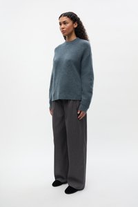 Maglione turchese testurizzato con scollo rotondo indossato sopra pantaloni a gamba larga grigi a righe sottili e ballerine nere, in piedi davanti a uno sfondo neutro.