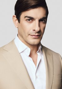 Mężczyzna w beżowej marynarce noszący białą koszulę, charakteryzującą się dopasowanym krojem, gładką fakturą oraz subtelnymi detalami, takimi jak akcent na klapie.