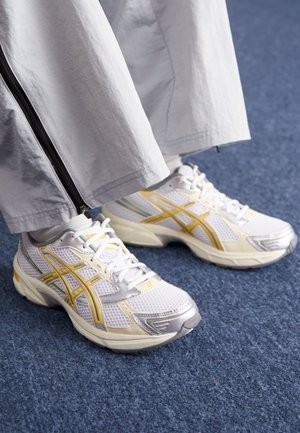 Personne portant des baskets blanches et jaunes avec un pantalon blanc debout sur un sol recouvert de moquette bleue.