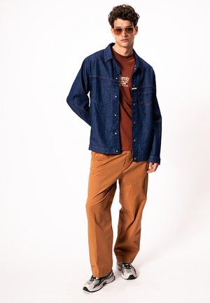 Mladý muž v hnedom slnečných okuliaroch, tmavomodrej denimovej bunde, hnedom tričku s potlačou, béžových nohaviciach a sivých teniskách stojaci na bielom pozadí.