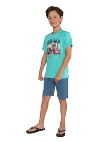 Een jongen draagt een turkooise t-shirt met korte mouwen, voorzien van een grafisch ontwerp, gecombineerd met blauwe shorts en zwarte slippers.