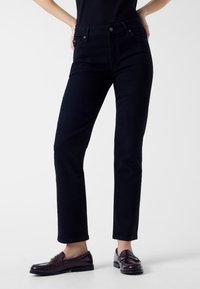 Jeans blu scuro a vestibilità slim, con tasche anteriori, chiusura con cerniera e bottone. Indossati con mocassini scuri.