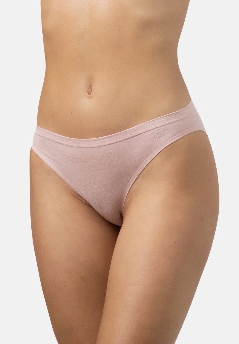 Femme portant un sous-vêtement en coton de style bikini rose clair sur un fond uni clair, avec la taille jusqu'au haut des cuisses visibles.