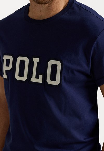 Tricou din bumbac bleu marin, având litere mari, brodate, "POLO" în alb cu o contur întunecat. Guler clasic, de tip crew și mâneci scurte.