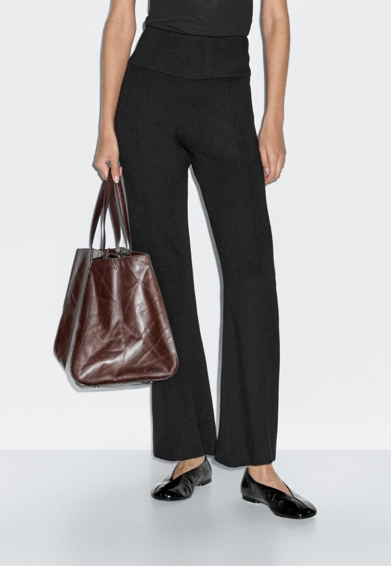 Zwarte flared broek met een hoge tailleband, gecombineerd met een gestructureerde bruine leren tote bag. Glanzende zwarte ballerina's maken de look compleet.