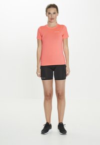 Short-sleeve athletic top in bright coral, paired with black shorts. Top has a fitted design, while shorts are mid-thigh length. Black sneakers complete the look.