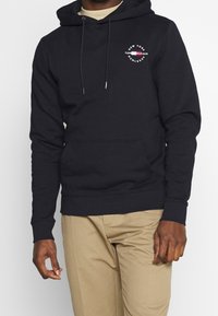 Sudadera azul marino confeccionada en una mezcla de algodón, que presenta un bolsillo frontal, cordones ajustables y detalles de logotipo en el pecho.