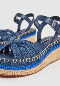 Tumedate sininad koos punutud tekstiilist rihmadega, tekstureeritud espadrille-stiilis tallaga ja kontrastsete siniste aktsentkihtidega. Klambriga kinnituse detail.