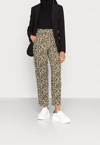 Olivengrüne Druckhose mit schwarz-weißen abstrakten Mustern, schmal zulaufendes Design, hohe Taille und Seitentaschen, kombiniert mit weißen Sneakers.