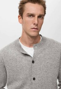 Cárdigan gris de punto con cuello, que presenta tres botones negros, llevado sobre una camiseta blanca. La textura parece suave y acogedora.