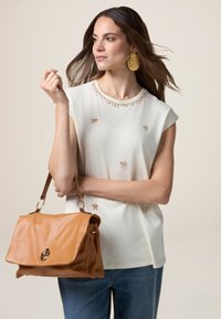 Weißes Kurzarmoberteil mit perlenbesetzten Blumenakzenten, kombiniert mit einer braunen Lederhandtasche, die ein strukturiertes Design und goldene Hardware aufweist.