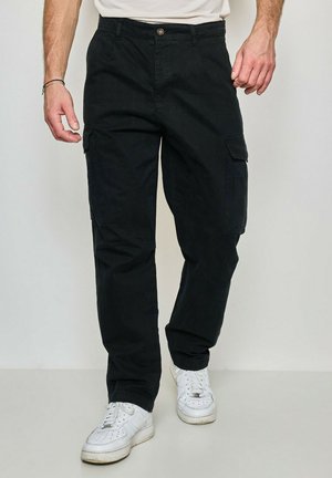Homme portant un pantalon cargo noir avec des poches latérales et des baskets blanches, debout devant un fond clair uni.