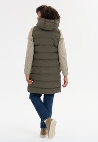 Khaki Steppweste mit Kapuze, strukturierten grauen Ärmeln und knielangem Schnitt. Kombiniert mit blauen Jeans und beigen Stiefeln bis zum Knöchel.