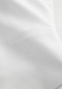 Gros plan d'une manche de veste en tissu blanc montrant un détail de poche soigneusement cousu sur un matériau à texture lisse.