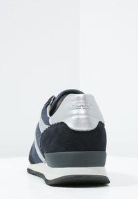 Mörkblå och silver sneakers sedda bakifrån, med textilstruktur, ett vitt rand och en svart sula på en vit yta.