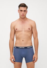 Férfi kék boxer alsónadrág fekete derékrésszel, amelyen fehér logó található. Simább anyagból készült, testhez álló fazonú és közepes hosszúságú.