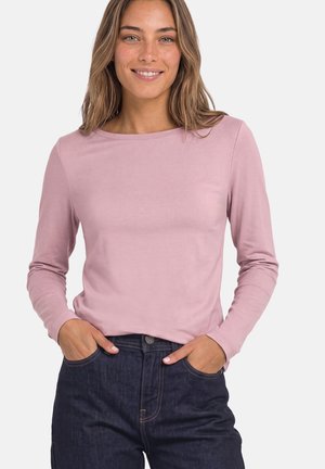 Femme aux cheveux châtain clair sourit, portant une chemise rose clair à manches longues et un jean bleu foncé, les mains dans les poches.