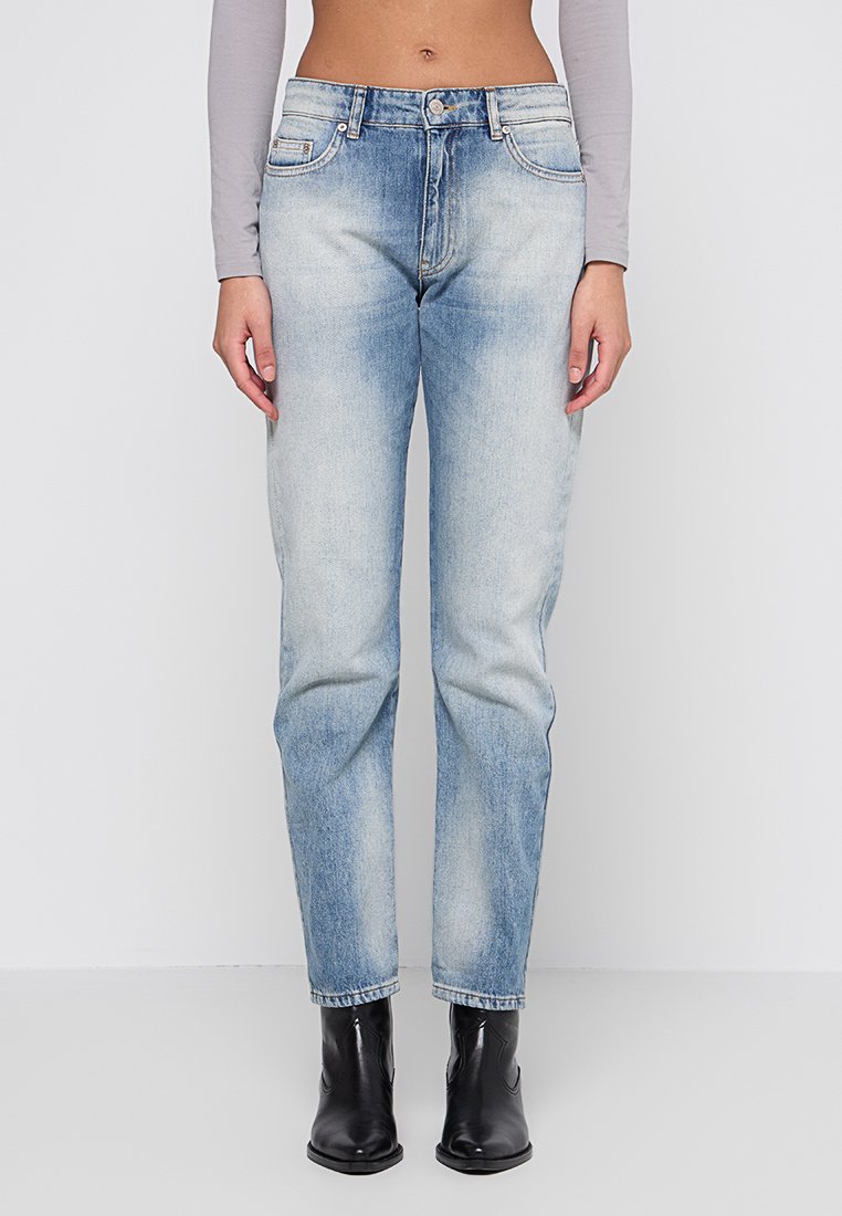 Persona con jeans de tiro alto en un azul claro y lavado desgastado, top corto de manga larga gris y botas negras de tobillo, posando frente a un fondo blanco.
