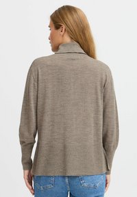 Pull en maille marron à manches longues avec un col montant et une coupe décontractée, présentant une texture côtelée et un ourlet fendu à l'arrière, porté avec un jean bleu.