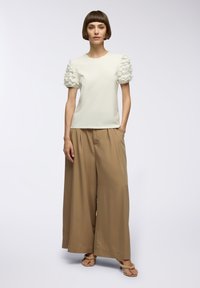 Top bianco con maniche a volant, abbinato a pantaloni beige a gamba larga. Il modello è in piedi su uno sfondo neutro, con le mani nelle tasche. Sandali piatti.