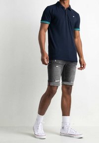 Polo navy con polsini a righe verdi e bianche, abbinato a pantaloncini di denim grigio usurati e sneakers bianche alte.