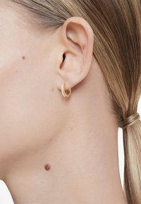 Gold hoop earring with a sparkling row of clear stones along the front edge, worn on a model's ear. Smooth material and circular design.