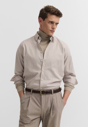 Junger Mann trägt ein beiges kariertes Hemd über einem beigen Rollkragenpullover, beigen Hosen und einen braunen geflochtenen Gürtel, steht mit den Händen in den Taschen.