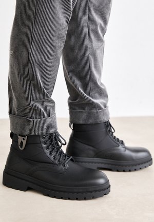 Bottes en cuir noir à lacets portées avec un pantalon gris à fines rayures retroussé, sur un sol clair devant un fond blanc.