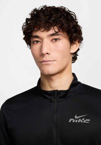 Musta urheilutakki vetoketjullisella kauluksella, jossa rinnan kohdalla harmaa Nike-logo. Sileä pinta ja istuva muotoilu.