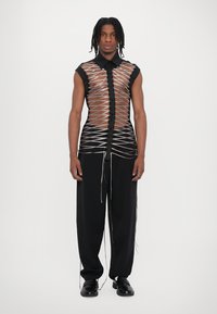 Chemise noire et transparente avec un col, présentant des détails en rubans croisés et une patte de boutonnage sur le devant, accompagnée de pantalons noirs.