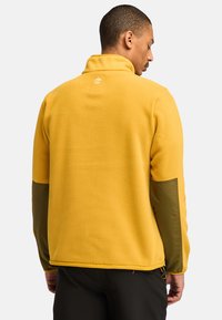 Gelbe Fleecejacke mit olivgrünen Ärmeln, hohem Kragen und kleinem Logo auf der Rückseite. Verfügt über elastische Bündchen und einen mit Reißverschluss versehenen Saum.