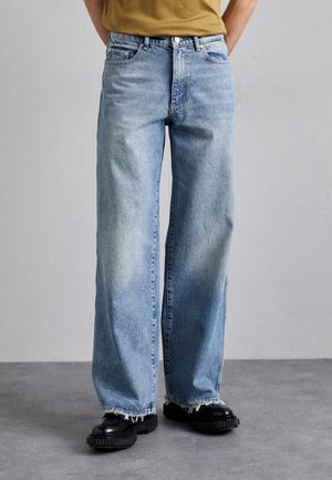 Person trägt hellblaue, weit geschnittene Jeans mit ausgefransten Säumen und schwarzen klobigen Schuhen und steht auf einem grauen Boden vor einer schlichten Wand.