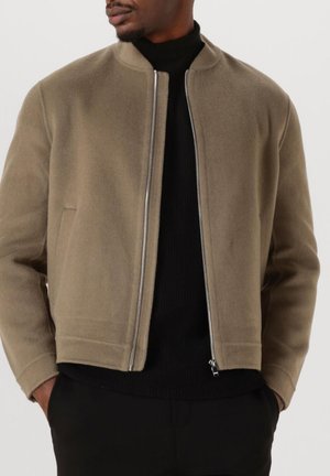 Mann in einer hellbraunen Zip-Jacke über einem schwarzen Rollkragenpullover, mit den Händen in den Taschen einer schwarzen Hose.