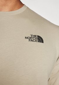 Detailní záběr na béžové tričko v oblasti hrudníku s černým logem "The North Face" na levé straně.
