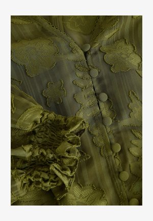 Bluzka w oliwkowej zieleni z haftem w kwiatowe wzory, teksturowana tkanina, dekoracyjne guziki oraz falbaniaste rękawy, które podkreślają skomplikowane detale haftu.