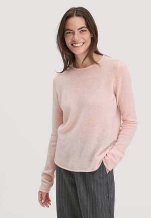 Jonge vrouw die glimlacht, gekleed in een lichtroze sweater met lange mouwen en donkergrijze broek met streepjes, staand met één hand in haar zak.