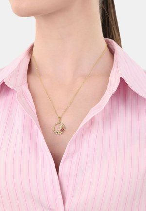 Collana d'oro con un ciondolo circolare decorato con un motivo floreale e un accento rosso, indossata sopra una camicia a righe rosa chiaro.