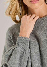 Blonde Frau trägt einen grauen Strickpullover mit gepufften Ärmeln und langen, hängenden Ohrringen, Hand nahe am Hals, Lippen sichtbar.