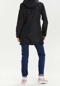 Parka impermeabile nera con cappuccio, vita aderente e maniche lunghe. Abbinata a jeans blu e sneakers di colore chiaro.