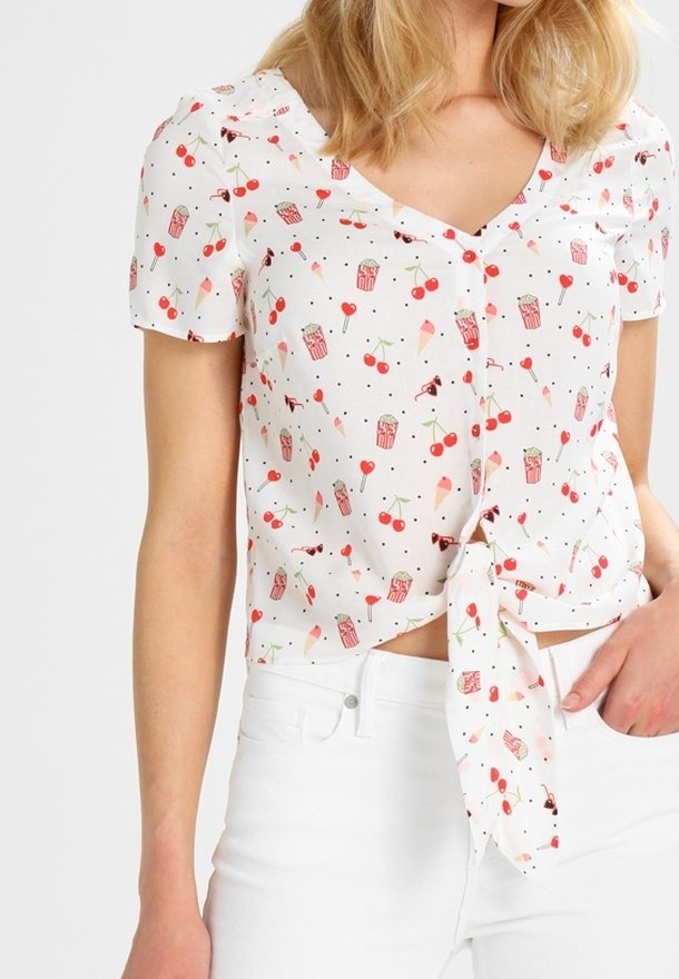 Femme portant un chemisier blanc boutonné avec des imprimés de cerises rouges, de glace et de popcorn noué à la taille, associé à un pantalon blanc.