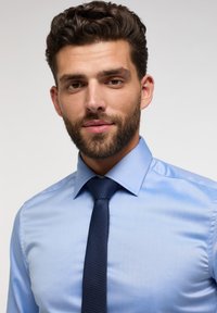 Chemise de couleur bleu clair avec une texture lisse et un col pointu, associée à une cravate bleu foncé texturée, centrée sur la zone du cou.