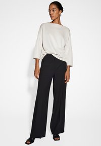 Frau mit einem Dutt, die ein lockeres weißes Oberteil, weite schwarze Hosen und offene schwarze High Heels trägt, steht vor einem weißen Hintergrund.