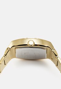 Guld-tone armbåndsur med rektangulært hus, metallisk finish, en teksturert rem og en sidekrone for justering av tid. Baksiden viser inngravede detaljer.