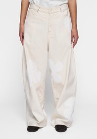 Pantalones de mezclilla beige de pierna ancha con grandes patrones florales blancos. Presentan bolsillos delanteros y un cierre de botón en la cintura.