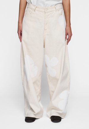 Pantaloni beige a gamba larga con grandi motivi floreali bianchi, realizzati in denim. Presentano tasche frontali e una chiusura a bottoni in vita.