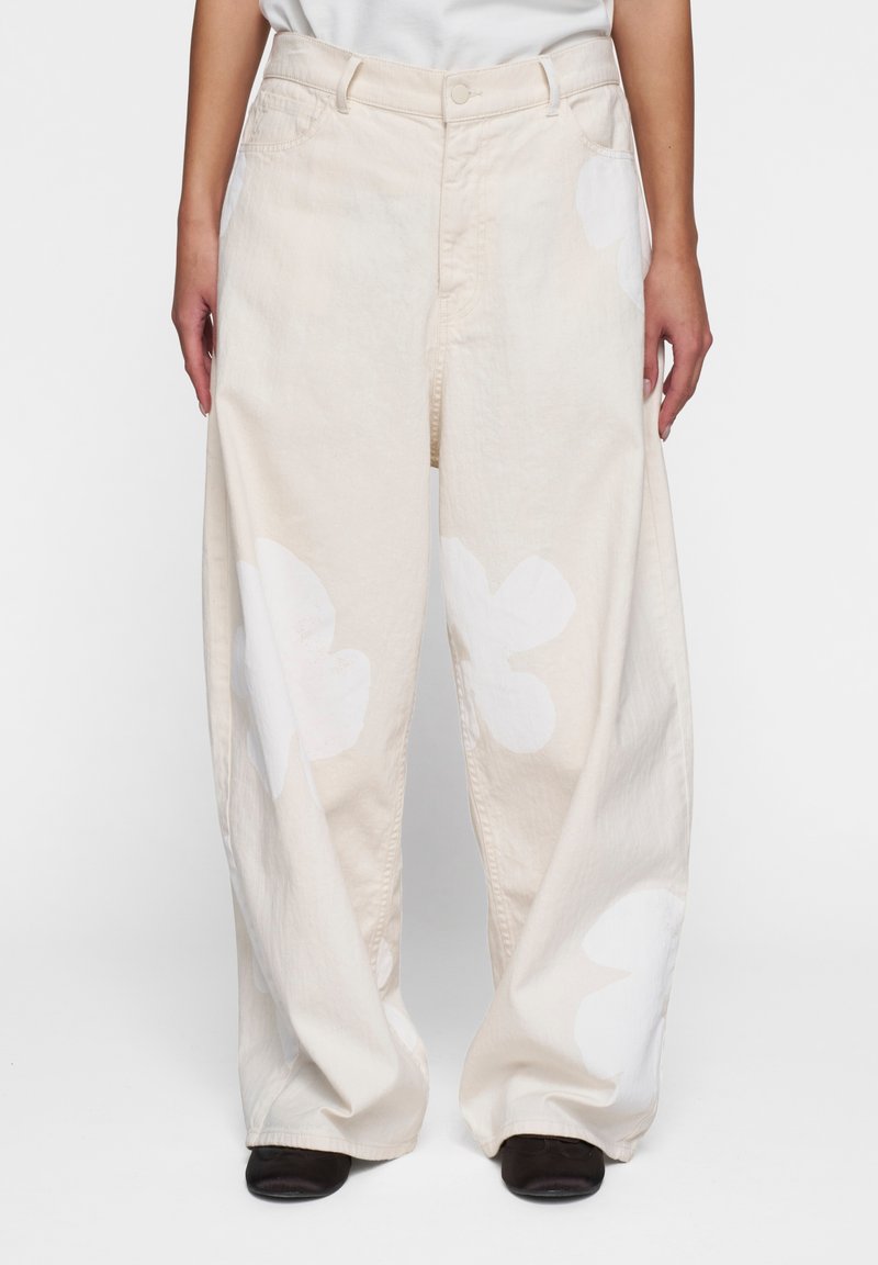 Pantalones de mezclilla beige de pierna ancha con grandes patrones florales blancos. Presentan bolsillos delanteros y un cierre de botón en la cintura.