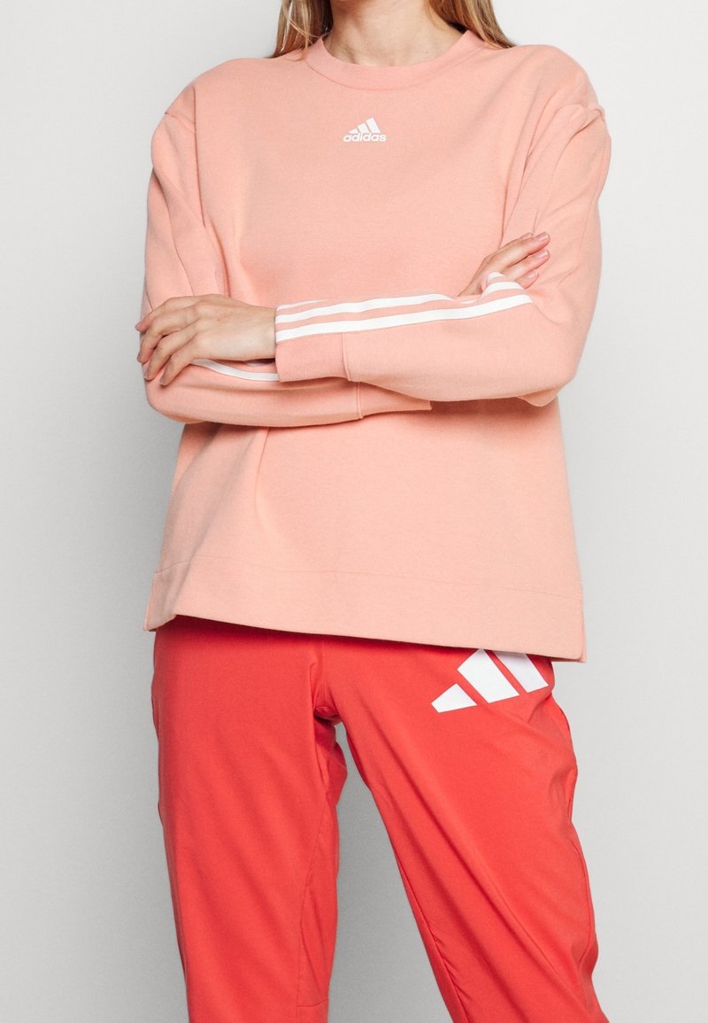 Personne portant un sweat-shirt Adidas rose clair avec des bandes blanches sur les manches et un pantalon rouge Adidas avec un logo blanc sur la partie supérieure de la cuisse.